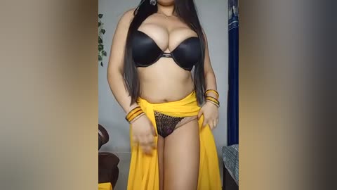 Media: A video of a curvy, light-skinned woman with long black hair, wearing a black bra, yellow wrap, and black lace panties, standing in a room with a brown chair and plant.