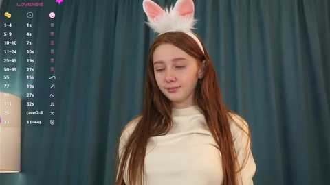 Media: Video of a young Caucasian woman with long red hair, wearing a white bunny headband, and a beige turtleneck sweater. Background features teal curtains and a digital overlay with a menu and statistics.