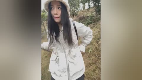 Media: A video of a young Asian woman with long black hair, wearing a white jacket and a beige hat, standing outdoors in a forested area with dry grass and trees in the background.