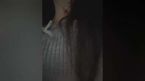 Media: A dimly lit, close-up video of a person's head and shoulders, partially obscured by darkness. The individual, likely a woman with long, dark hair, is wearing a grey, ribbed sweater. The image has a moody, introspective atmosphere.