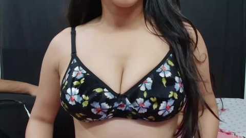 Media: Video of a woman with long, dark hair, wearing a black floral-patterned bra with white and blue flowers, revealing her medium-sized breasts. Background shows a dark curtain and a partially visible person.