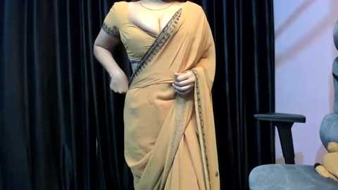 Media: Video of a fair-skinned woman in a mustard-yellow sari with a black border, wearing a black bra underneath. She stands against black curtains, with a teal chair and table in the background.