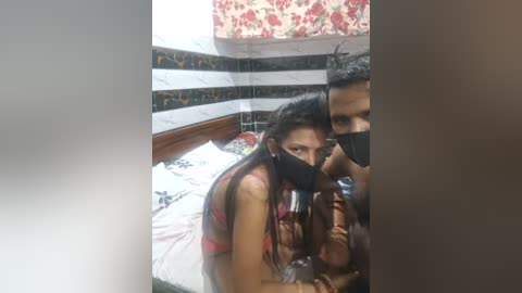 Media: Video of a young woman with long hair, wearing a pink bra and black mask, kneeling beside a man in a bed with floral-patterned walls.