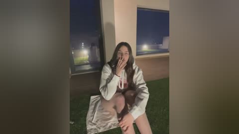 Media: Video of a young Asian woman with long black hair, wearing a white lab coat and shorts, sitting on a grassy lawn at night, partially obscured by a building, smoking a cigarette.