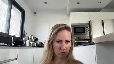 Media: Video of a blonde woman with a neutral expression in a modern kitchen with white cabinets, stainless steel appliances, and a window with a view of bare trees.