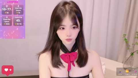 Media: Video of an East Asian woman with straight black hair and fair skin, wearing a black strapless top with a red bow, in a modern room with a white curtain.