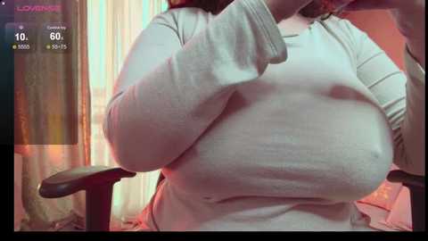 Media: Video of a plus-sized woman with large breasts, wearing a tight white long-sleeved top, sitting on a black chair in a dimly lit room with a pinkish glow.