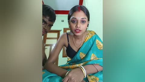 Media: Video of a young woman with dark skin and red dot on forehead, wearing a blue sari with golden patterns, sitting on a bed; a boy with short dark hair peeks from behind.