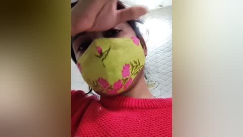 Media: Video of a person with light brown skin, wearing a bright yellow mask with pink floral patterns, covering the nose and mouth. The person has brown hair tied back, wearing a red knit sweater. The background shows a white wall with a quilted texture.