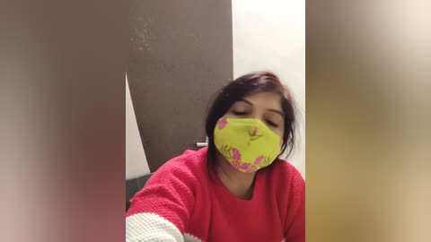 Media: Video of a woman with medium skin tone, wearing a bright green face mask with pink designs, red sweater, and white gloves, leaning against a beige wall, blurred background.