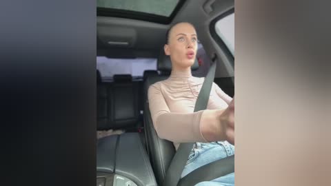 Media: Video of a blonde woman with fair skin, wearing a beige long-sleeve top and blue jeans, driving a car, looking focused, with a dark interior background.