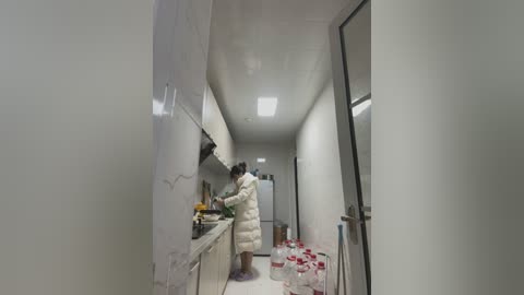 Media: Video of a narrow, modern, white-tiled kitchen with a woman in a white puffy coat, cooking, and plastic bottles scattered on the floor.