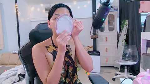 Media: Video of an Asian woman in a black tank top with a floral pattern, drinking from a glass, in a cluttered, dimly lit room with a white desk, chair, and string lights.