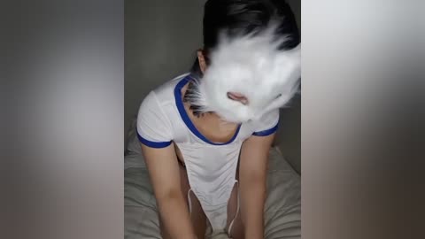 Media: Video of a person wearing a white, translucent mask with a blue trim, kneeling on a bed with beige sheets. The mask covers the face, obscuring features. The room is dimly lit, with soft shadows.