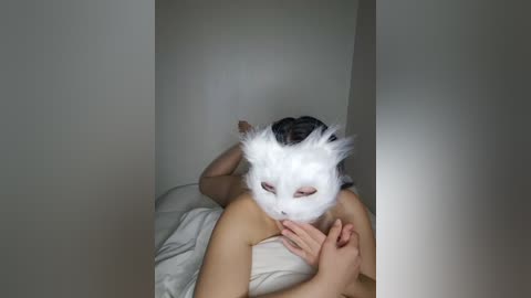 Media: A video depicts a person in a white cat mask, lying on a bed with white sheets, hands resting on their chest, creating a mysterious and enigmatic atmosphere.