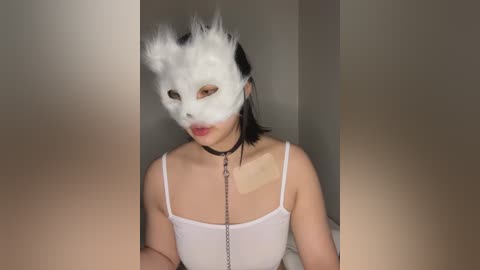 Media: Video of a young Asian woman with light skin and straight black hair, wearing a white feathered masquerade mask, a black choker, and a white spaghetti-strap top. She holds a chain leash in her right hand.