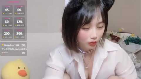 Media: A video of an East Asian woman with black bob cut hair, wearing a white shirt, black cat ears, and a choker, sitting indoors with a duck plush toy and a live streaming screen showing temperature and viewers.