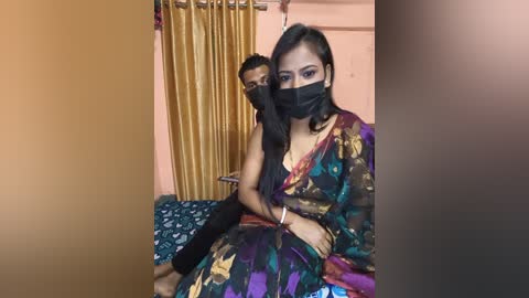 Media: A video shows a South Asian woman in a colorful sari with a mask, holding a masked man with a bandaged arm on a bed, in a modest room with yellow curtains and a green bedspread.