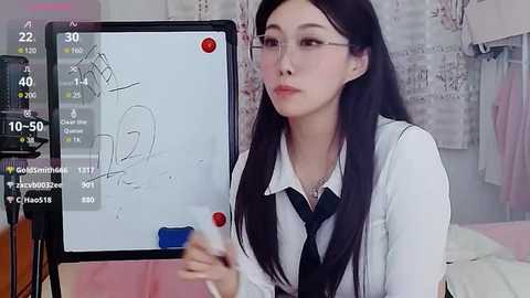 Media: A video of a young East Asian woman with long black hair, wearing glasses, a white shirt, and a black tie, standing in front of a whiteboard in a room with hanging clothes.