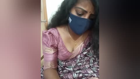 Media: Video of a South Asian woman with medium brown skin and long black hair, wearing a pink and white floral saree, blue face mask, and gold necklace, leaning against a door frame.