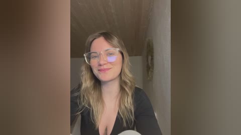 Media: Video of a smiling Caucasian woman with wavy blonde hair, wearing glasses, a black top, standing in a dimly lit hallway with wooden ceiling and beige walls.