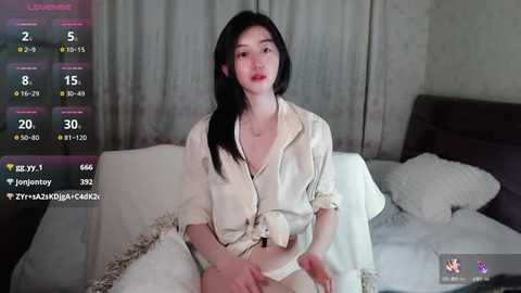 Media: Video of an East Asian woman with long black hair, wearing a satin cream nightgown, sitting on a bed with white linens.