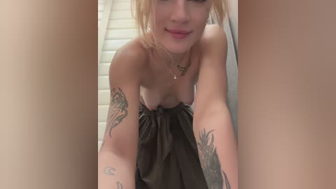 Media: Video of a topless woman with blonde hair and fair skin, wearing a black cloth, tattoos on her arms, in a bathroom with white tiles.