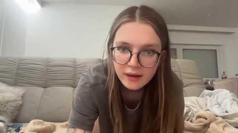 Media: Video of a young Caucasian woman with long, straight brown hair and glasses, lying on a beige couch in a cozy living room, surrounded by soft furnishings and a cluttered blanket.