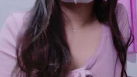 Media: Video of a woman with long, wavy brown hair, wearing a pink, low-cut, sheer blouse, and a white mask, with a small pendant necklace. The background is blurred in pink hues.