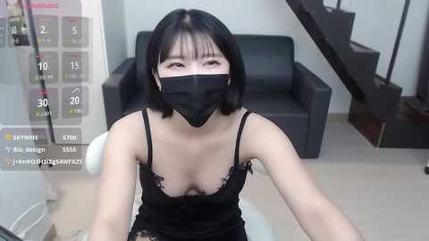 Media: A video of an East Asian woman with a short black bob haircut, wearing a black mask, black lace camisole, and black underwear, sitting in a modern living room with a black couch and stairs.