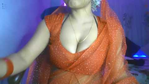 Media: A video of a fair-skinned woman in an orange saree with a plunging neckline, showcasing ample cleavage. She wears a blue bindi and a gold necklace, seated against a dimly lit background.