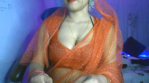 Media: A video of a light-skinned woman in a red, glittery saree and blouse, with a black choker and bracelets. She sits in a chair, hands on her lap, lips painted red, in a dimly lit room with purple and blue lighting.