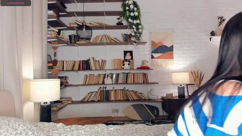 Media: Video of a cozy, modern living room with a woman in a blue and white striped shirt seated, facing a white brick wall with bookshelves and hanging plants.