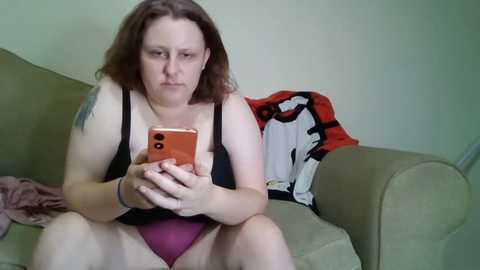 Media: Video of a plus-sized white woman with shoulder-length brown hair, wearing a black tank top and purple underwear, sitting on a beige couch, engrossed in her smartphone.