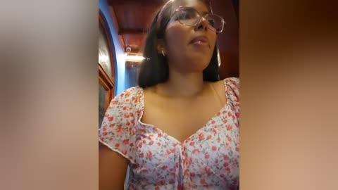 Media: Video of a young woman with medium skin tone, wearing glasses and a floral blouse, standing indoors in a warmly lit room with wooden paneling.