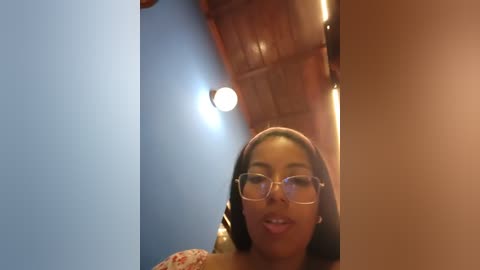 Media: A video of a young Black woman with medium brown skin and long black hair, wearing glasses, smiling. She is in a room with wooden furniture and a blue wall.