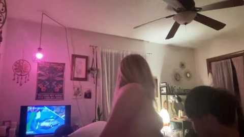 Media: Video of a dimly lit bedroom with a woman with long blonde hair, wearing a white shirt, playing a video game on a TV, while a man with short dark hair sits beside her.