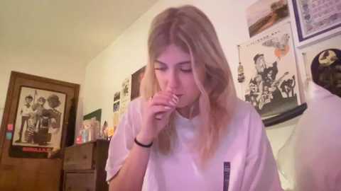 Media: Video of a young woman with long, straight, blonde hair, wearing a white T-shirt, eating a cookie while sitting in a cluttered bedroom with posters and a wooden dresser.