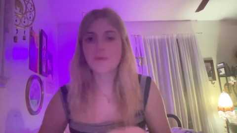 Media: A video of a young woman with long blonde hair, wearing a black and white striped top, standing in a dimly lit room with purple lighting, framed by wall hangings and a lamp.