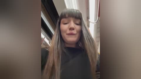 Media: A video captures a woman with straight, shoulder-length, gray-brown hair, wearing a black t-shirt, smiling with closed eyes, standing in a subway car with fluorescent lights and blurred passengers in the background.