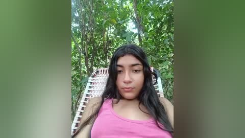 Media: A video of a young Latina woman with long black hair, wearing a pink tank top, reclining on a wicker chair in a lush, green forest.