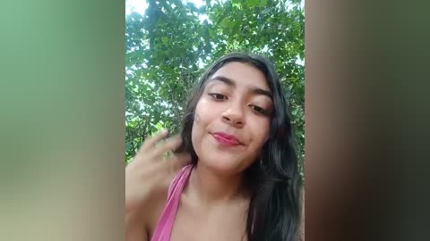 Media: Video of a young Latina woman with long black hair, light brown skin, and a medium build, wearing a pink tank top, smiling and looking at the camera against a green, leafy background.