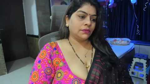 Media: Video of a South Asian woman with medium brown skin, wearing a vibrant pink sari with floral designs and a black dupatta. She has long, wavy black hair and is seated indoors, with a table and decorations in the background.