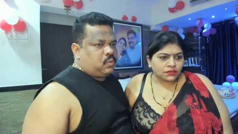 Media: Video of an overweight man with a mustache in a black sleeveless shirt and a woman in a red and black saree, standing in a brightly decorated room with a TV and balloons.