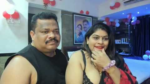 Media: Video of an Indian couple, a middle-aged man in a black sleeveless shirt and a woman with a red saree, holding hands in a brightly lit room with red balloons, framed photo, and a pool table.