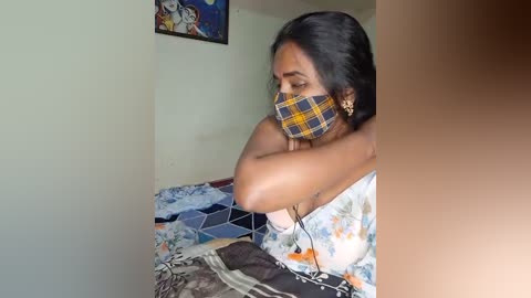 Media: Video of a South Asian woman with medium-dark skin, wearing a floral dress, black mask, and plaid headband, seated on a bed with a blue quilt, in a dimly lit room with a framed painting on the wall.