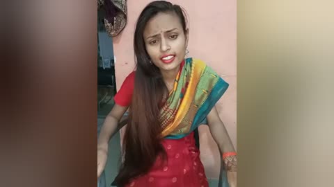 Media: Video of a South Asian woman with long brown hair, wearing a red sari with a green and yellow dupatta, sitting on a green floor, distressed expression, indoors.