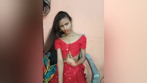 Media: Video of a young South Asian woman with long dark hair, wearing a red, off-shoulder crop top and matching skirt. She is seated on a blue plastic chair against a peach wall.