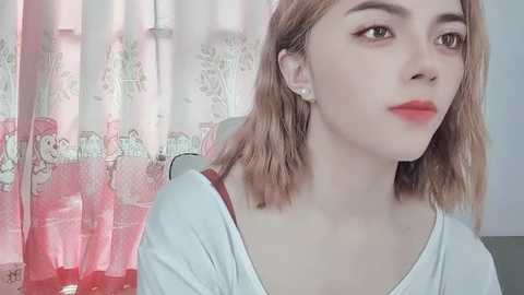 Media: Video of a young, fair-skinned Asian woman with shoulder-length, wavy, light brown hair, wearing a white top and a subtle red lip. She sits against a backdrop of pink floral curtains.