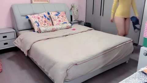 Media: Video of a cozy bedroom with light pink walls, a gray bed with beige bedding, floral pillows, a yellow shirt, and a woman's leg visible.